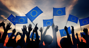 Hands raised against a sunset with silhouettes of various people displayed; many of the people are holding EU flags in the air