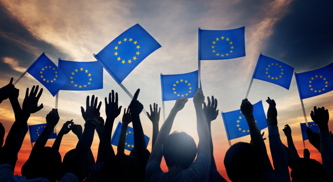 Hands raised against a sunset with silhouettes of various people displayed; many of the people are holding EU flags in the air